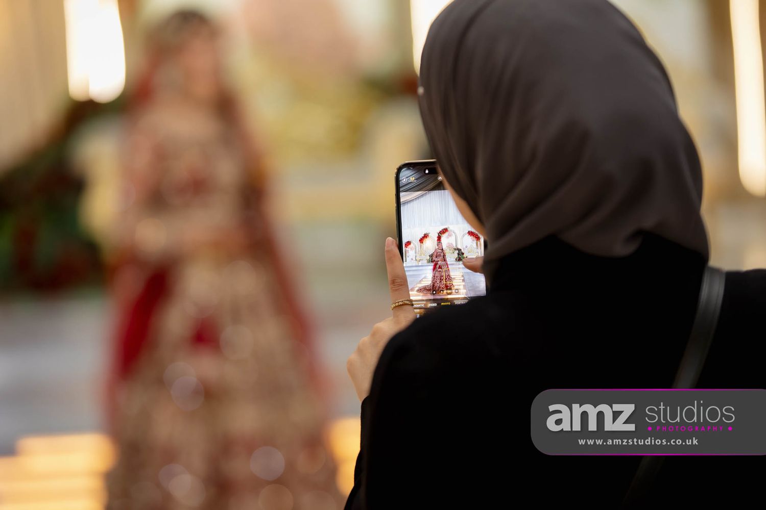 Woman in hijab photographs a bride in red bridal lehenga on smartphone at a wedding venue.