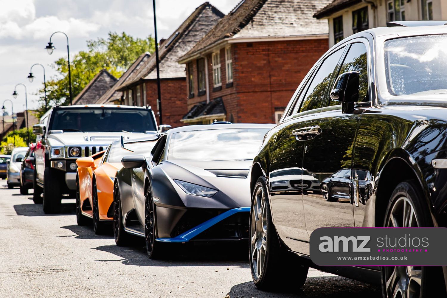 A row of luxury and exotic cars including a Lamborghini parked on a sunny suburban street.