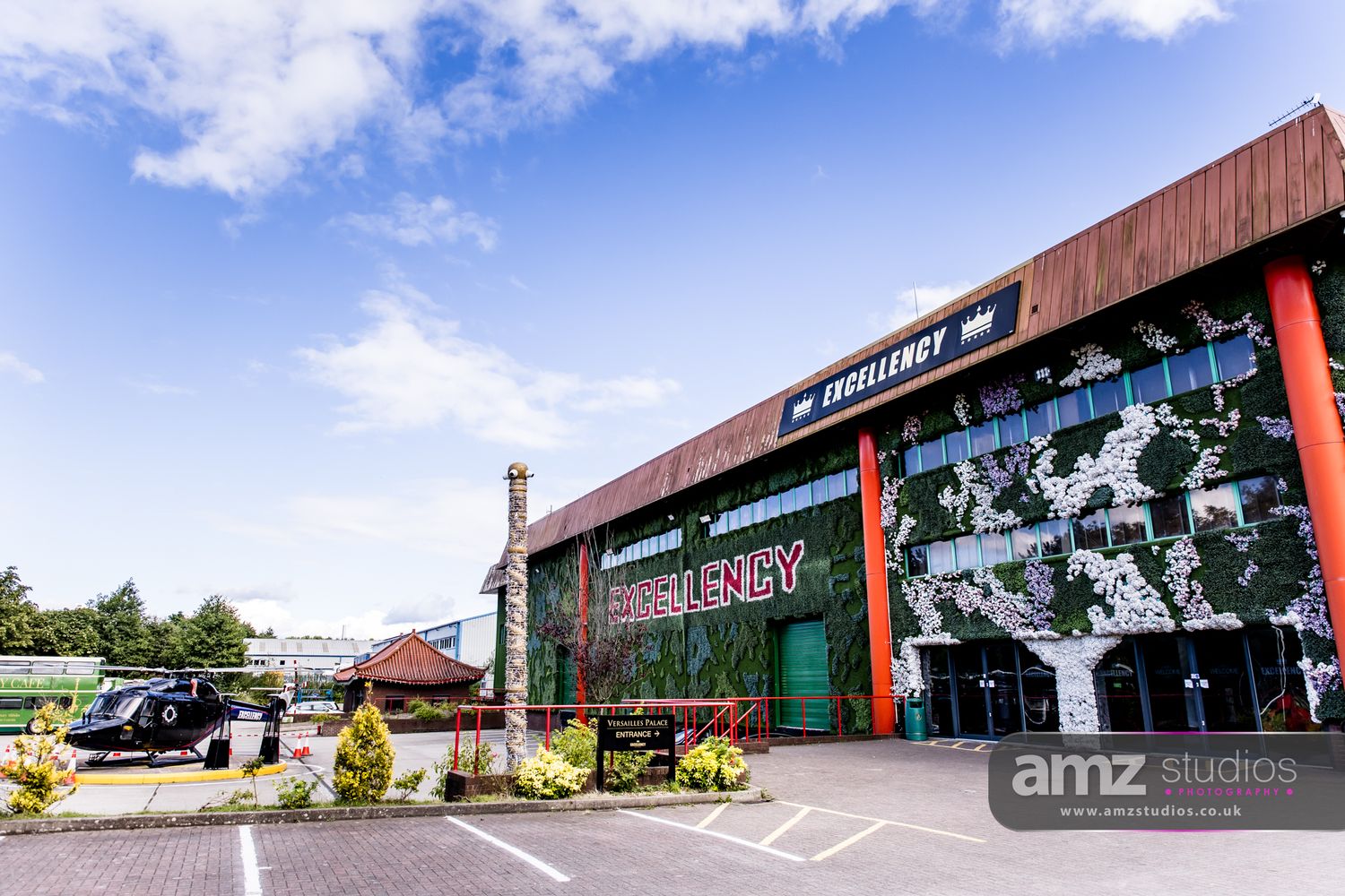 Modern commercial building with green wall facade, helicopter parked outside, blue sky backdrop, AMZ Studios watermark.