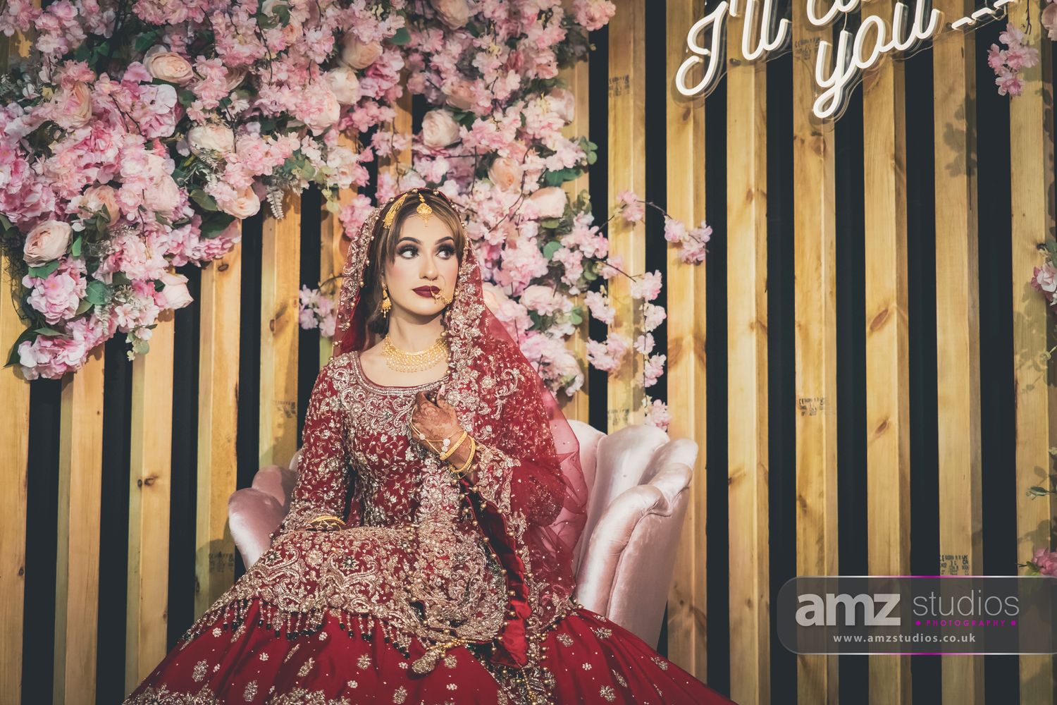 Bride in red bridal lehenga with gold embroidery poses before pink floral backdrop at South Asian wedding.