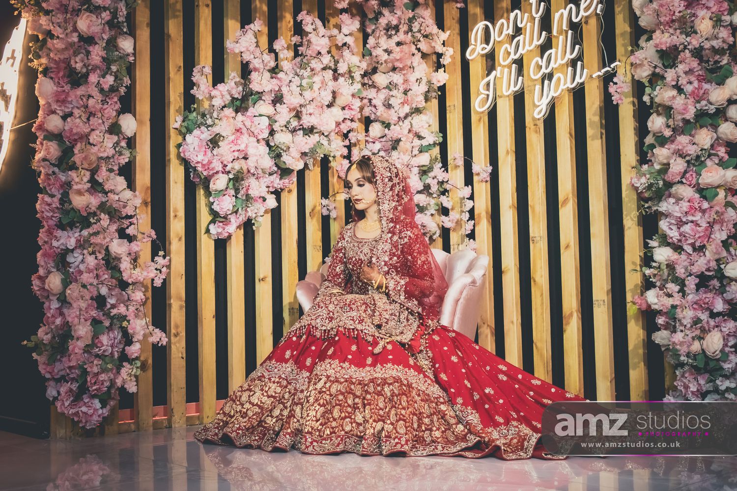 Bride in red and gold bridal lehenga poses gracefully before floral backdrop with neon sign at AMZ Studios.