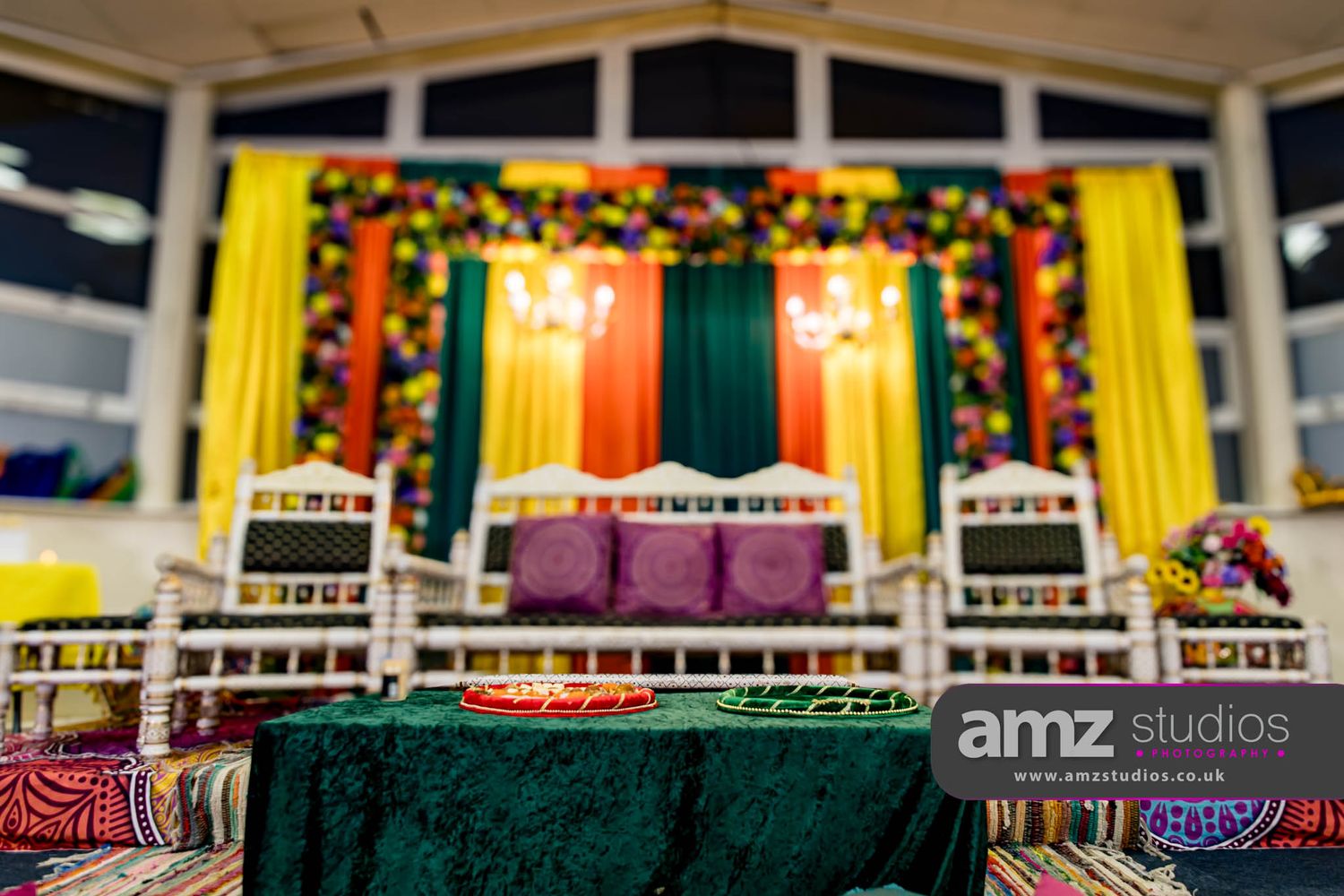 Colorful South Asian wedding stage with floral backdrop, elegant white chairs, and green draped table at a decorated venue.