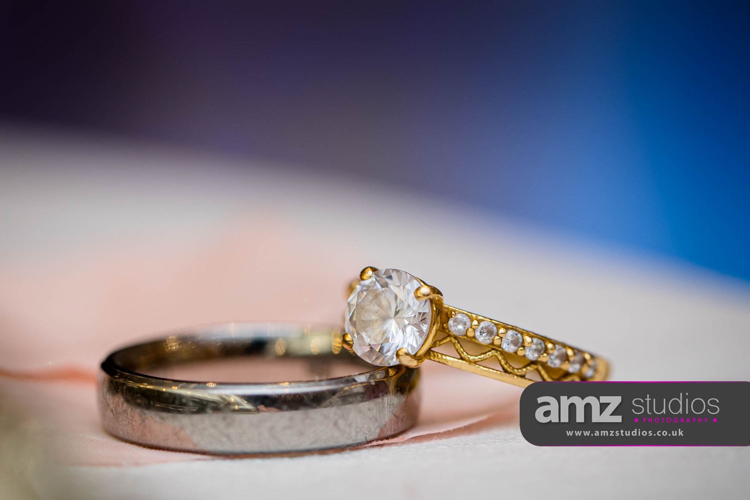 Gold diamond engagement ring and silver wedding band displayed together on a soft surface by AMZ Studios.