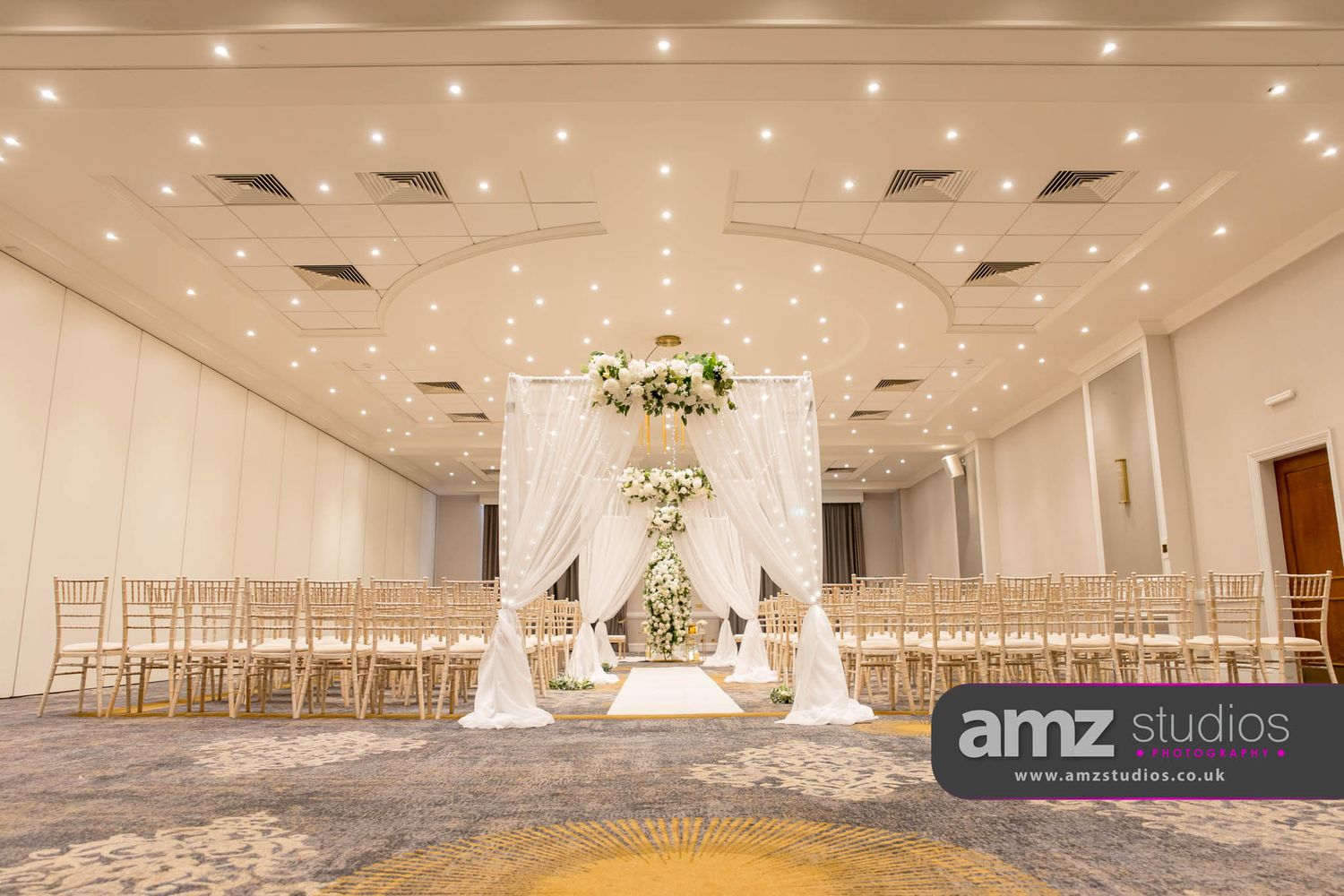 Elegant wedding ceremony setup with white draping, floral arch, gold chiavari chairs, and soft lighting in a grand ballroom.