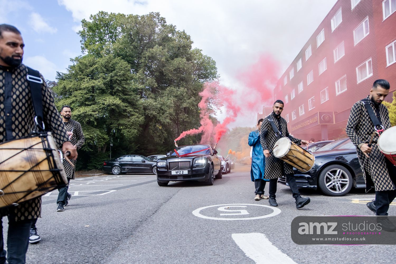 Men in traditional attire play dhol drums beside a decorated car with red smoke flares during a wedding baraat procession.