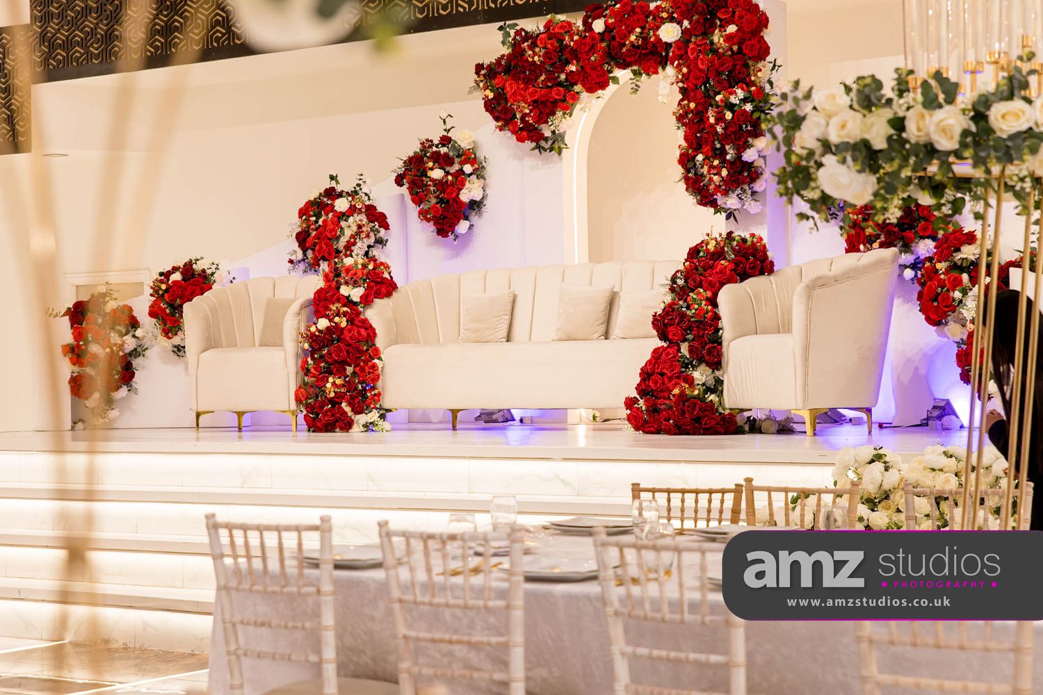 Elegant wedding stage with white sofas, red rose floral arrangements, and chiavari chairs at a luxury venue.
