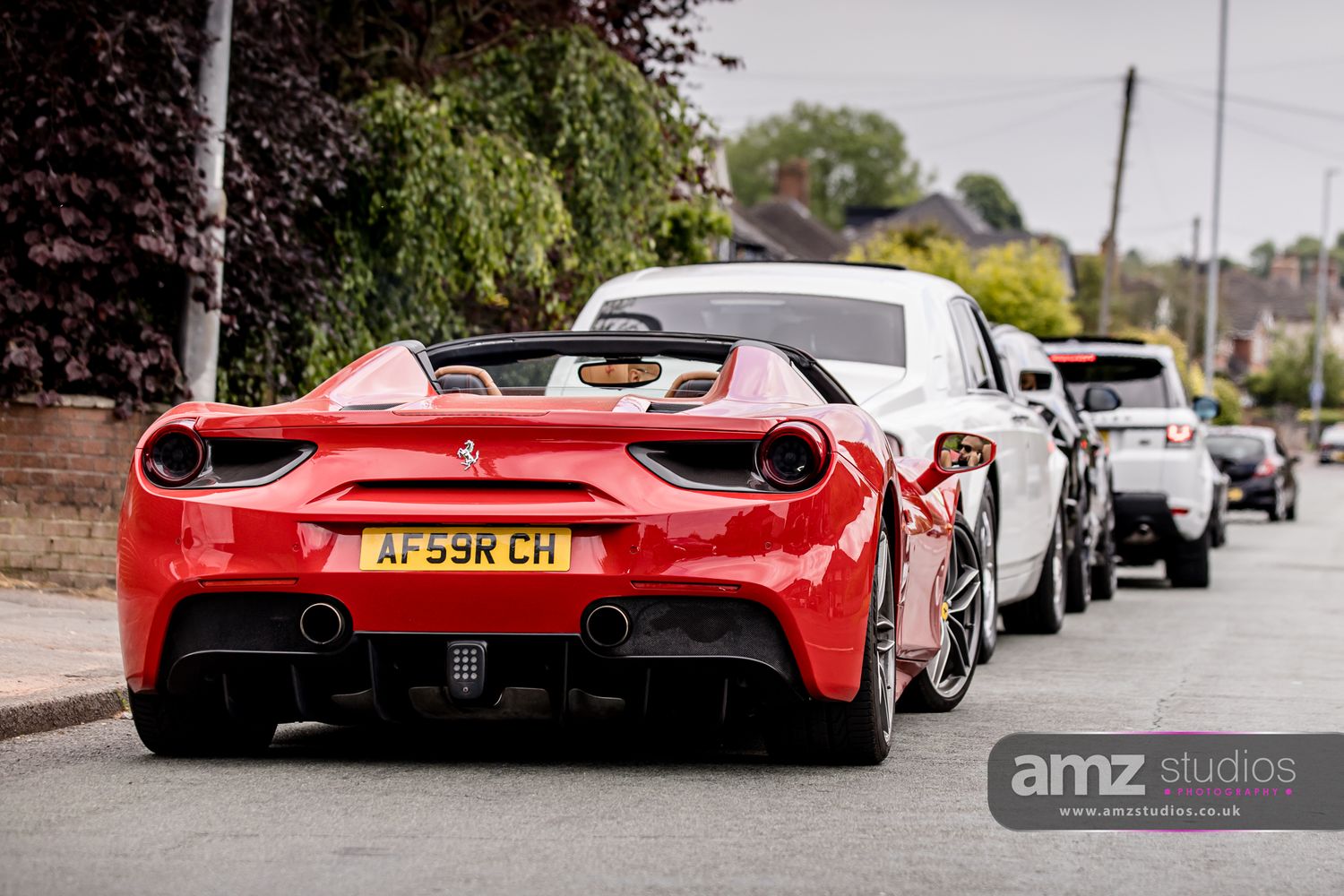 Red Ferrari 488 Spider with UK plate AF69RCH parked on a tree-lined street with white supercars behind.