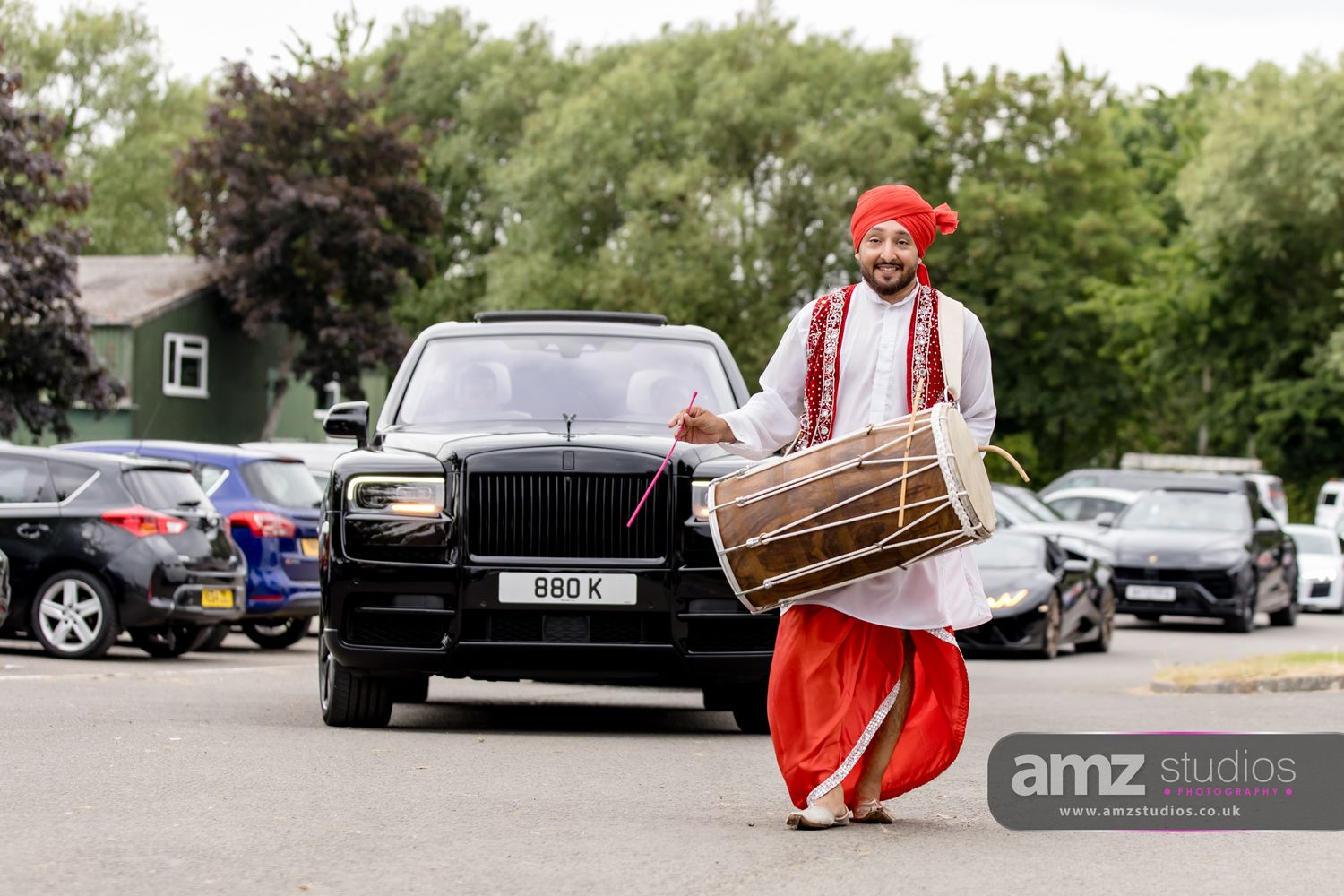 A Sikh dhol drummer in traditional red attire performs in front of a black Rolls-Royce at an outdoor wedding celebration.