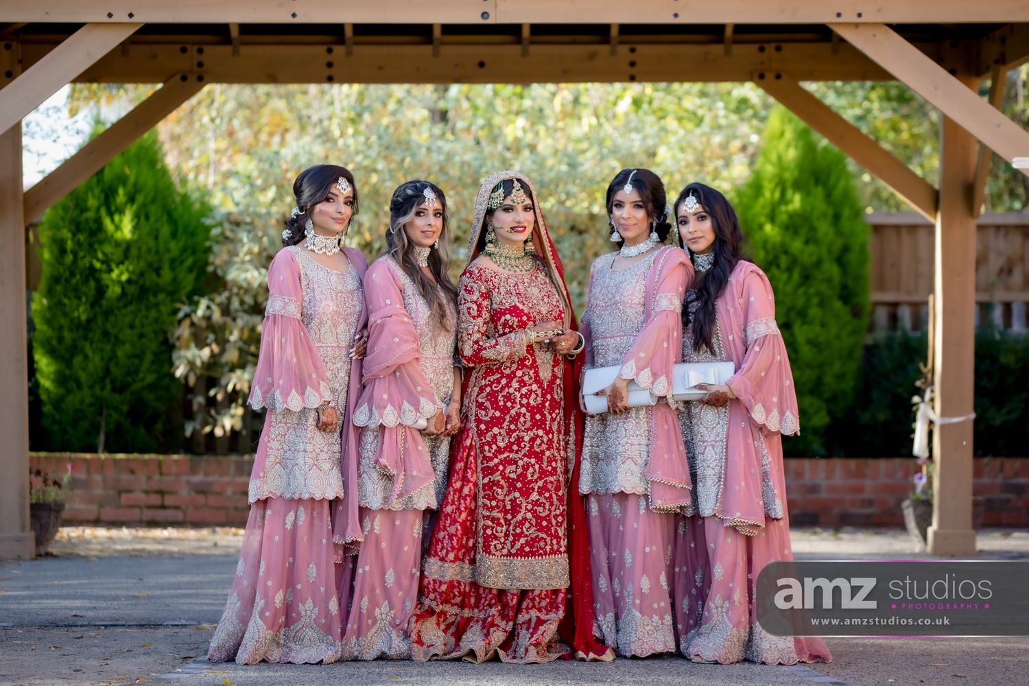 Bride in red lehenga poses with four bridesmaids in pink sharara suits at a South Asian wedding ceremony.