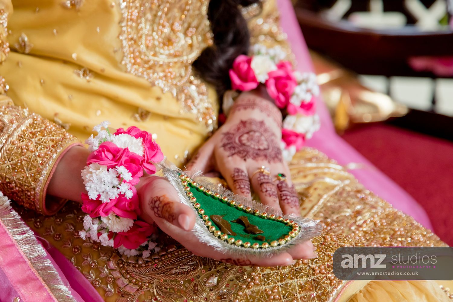 Bride's hands adorned with intricate mehndi henna, floral wrist jewelry, holding a green accessory in golden bridal attire.
