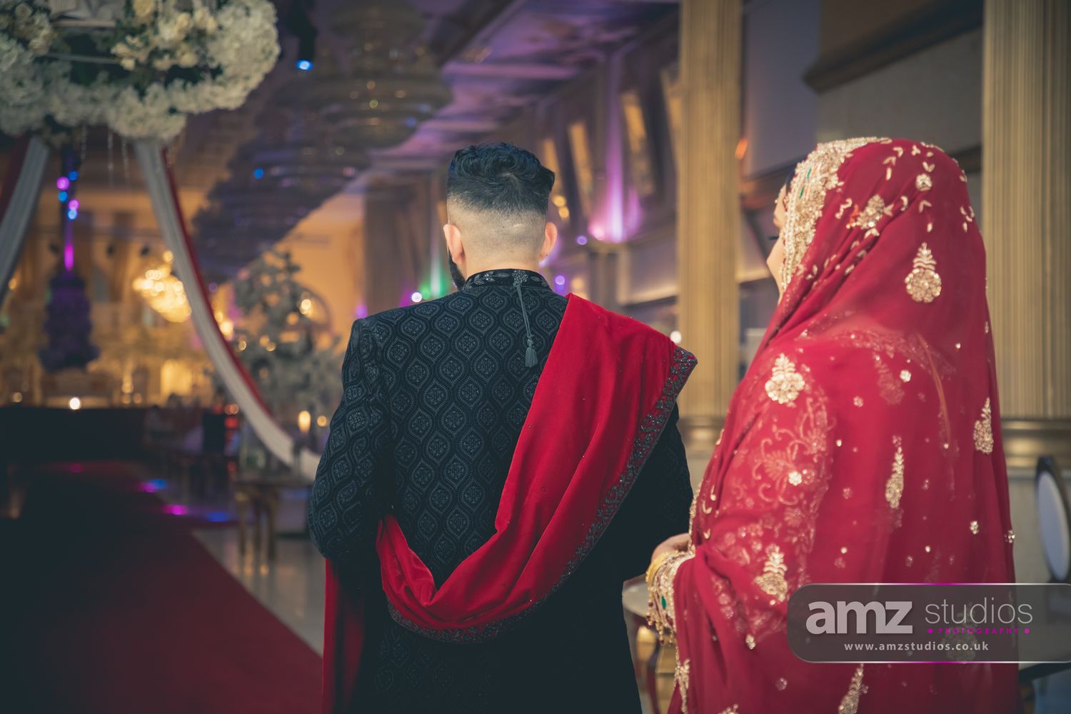 South Asian bride and groom in traditional red and black wedding attire walking together at an elegant wedding venue.