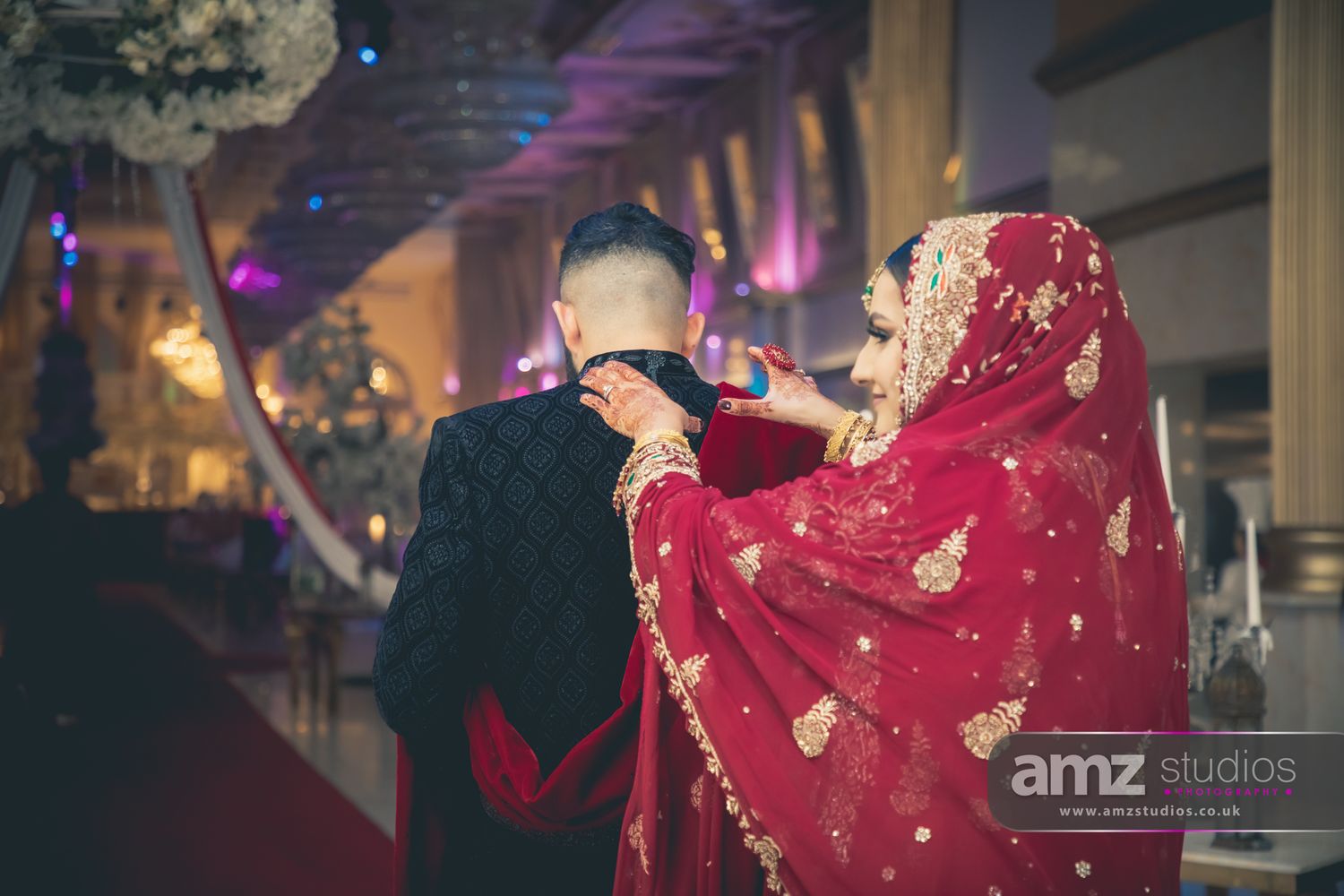 A bride in red dupatta adjusts groom's collar at a traditional South Asian wedding ceremony in an elegant venue.