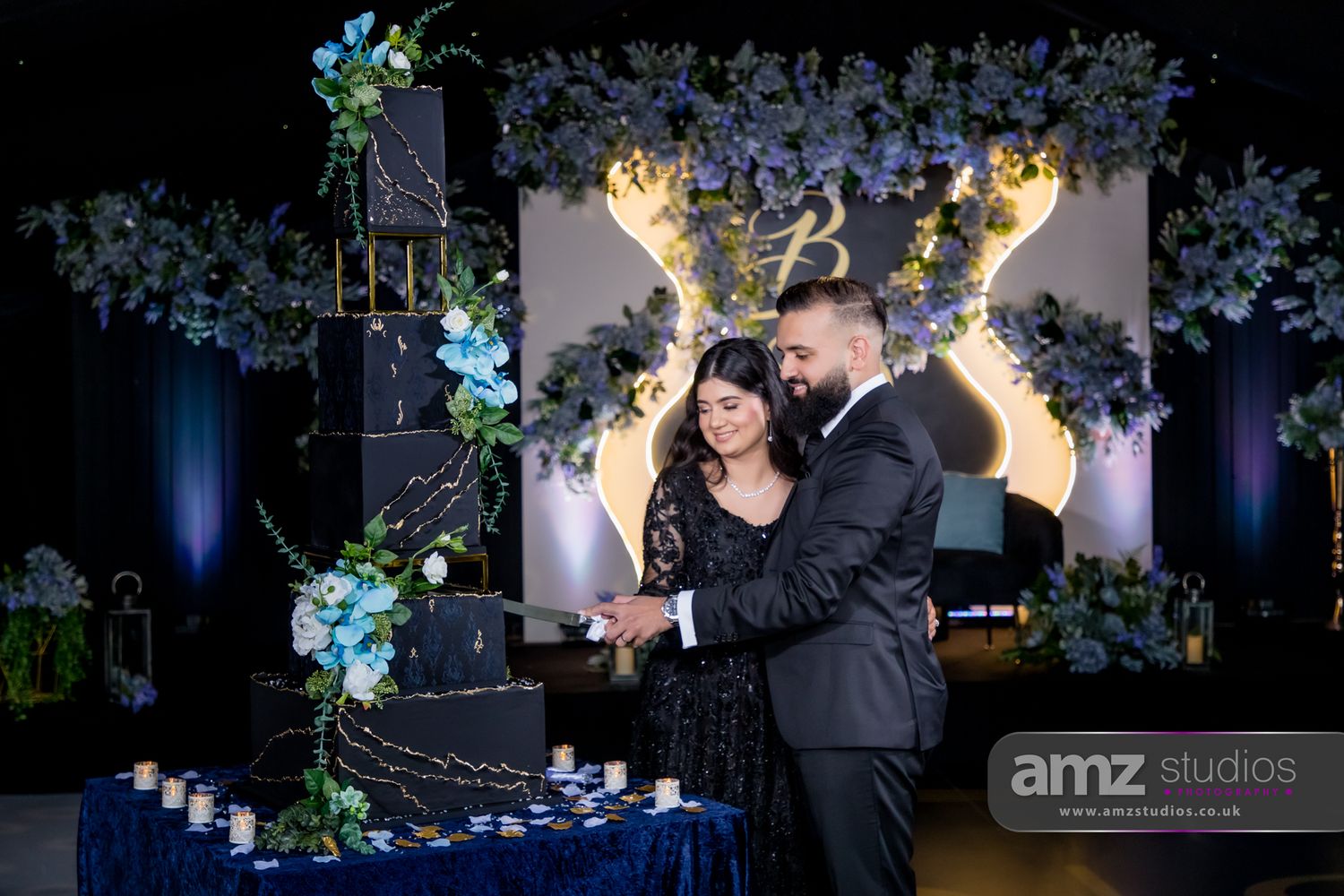 Couple cutting a black wedding cake with blue floral decor and glowing monogram backdrop at elegant reception.