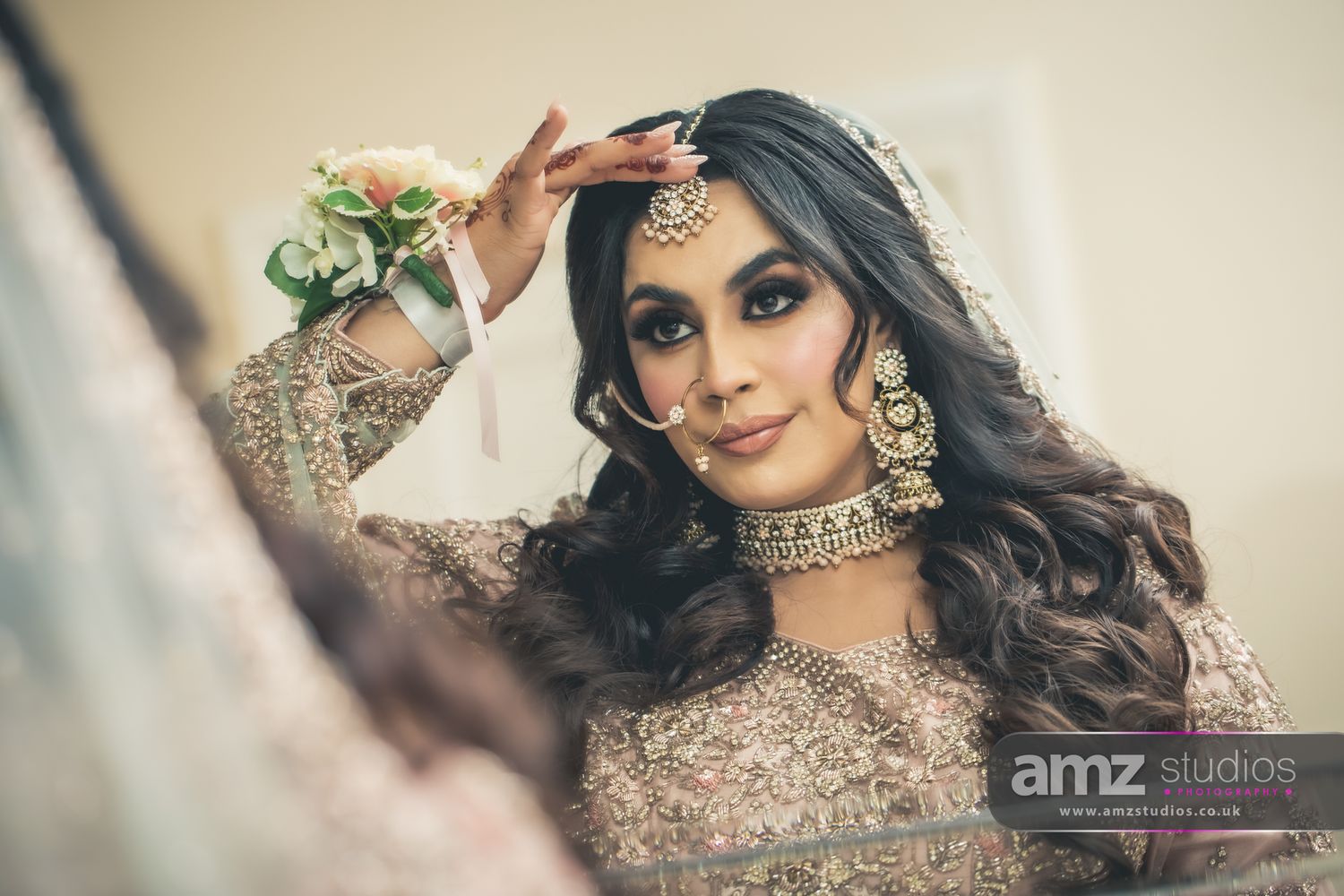 South Asian bride in ornate gold jewelry and embroidered lehenga poses elegantly during wedding preparations.