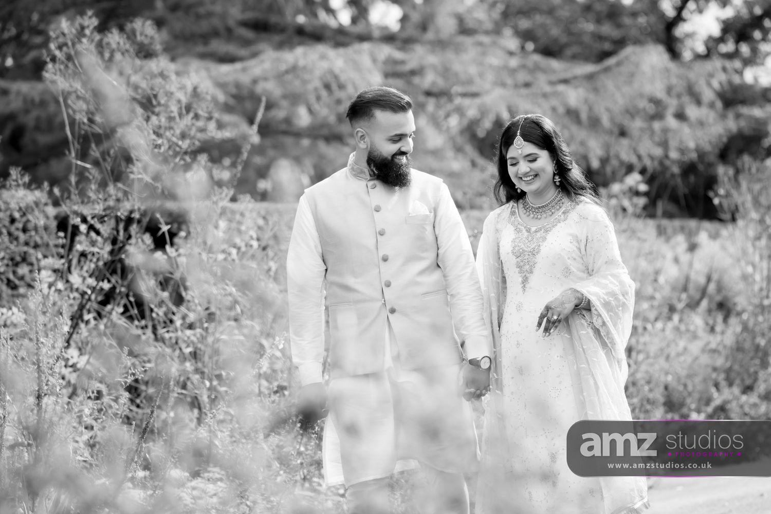 Black and white photo of a couple in traditional South Asian attire holding hands and smiling outdoors.