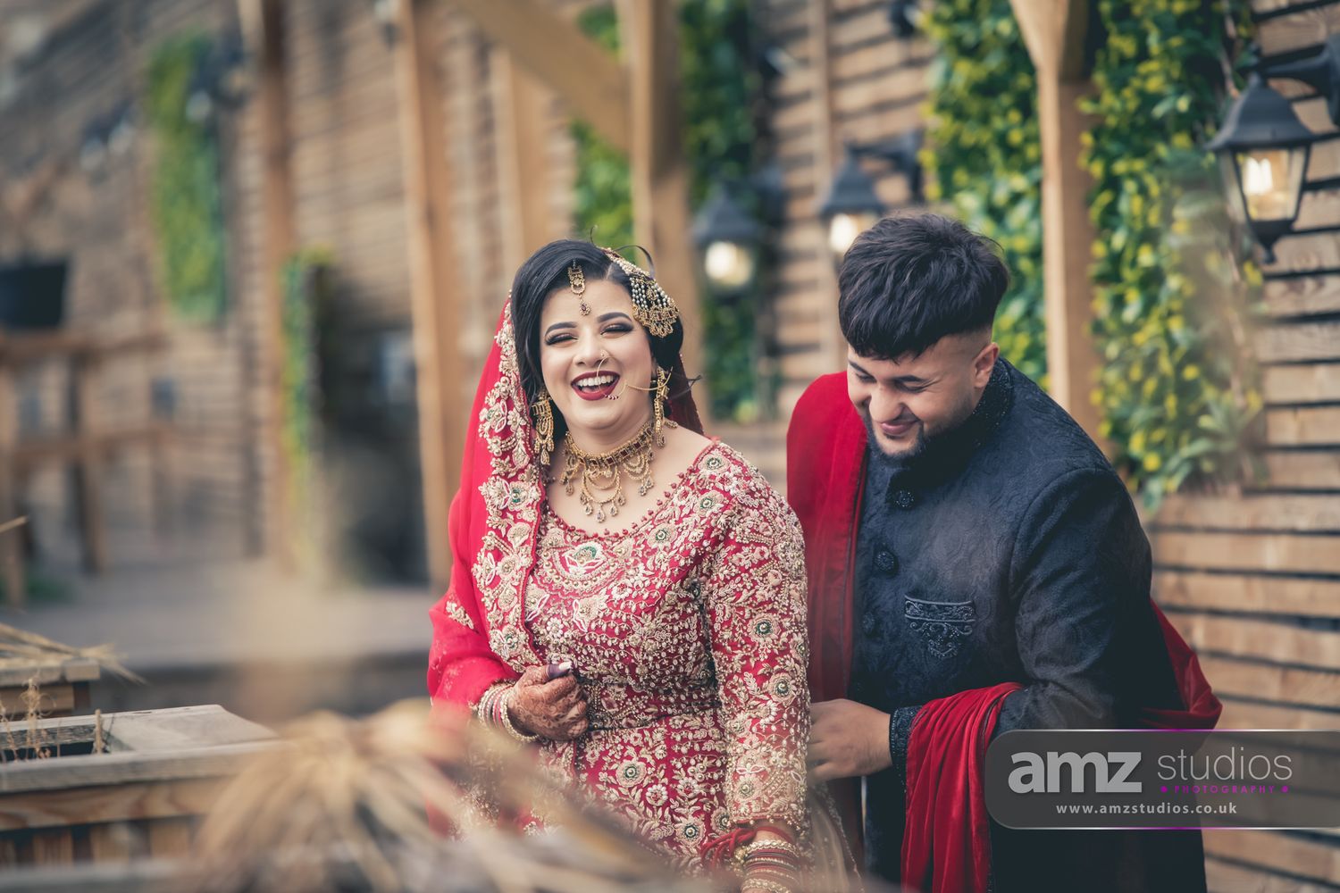 A smiling South Asian bride in red bridal attire poses with groom in black sherwani at an outdoor wedding venue.