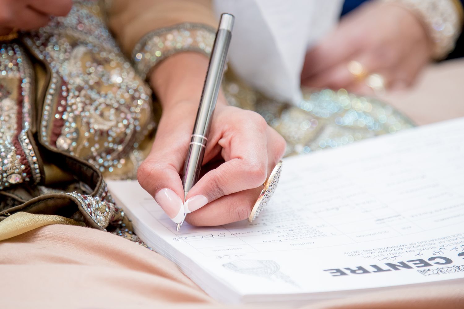 Close-up of a bride in embroidered attire signing a wedding document with a silver pen.