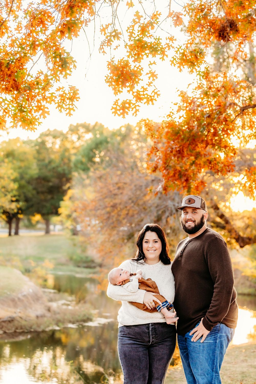 A couple poses with their newborn baby outdoors in a park during autumn, surrounded by vibrant orange and yellow foliage.