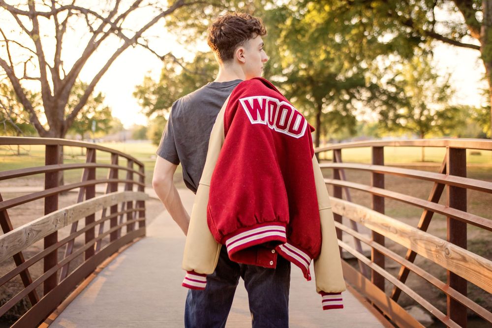 Teen boy holding red varsity letterman jacket on wooden bridge at golden hour sunset.