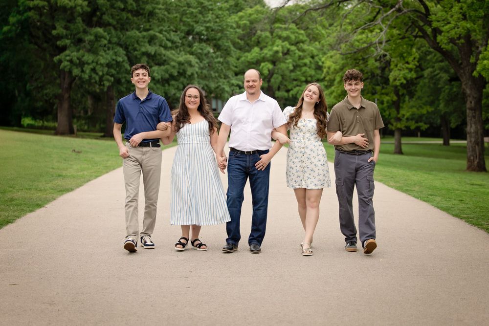 Happy family of five smiling and walking together on a park path surrounded by lush green trees.
