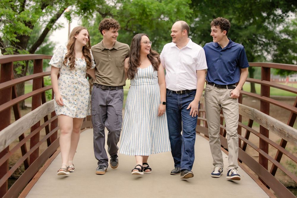 Family of five smiling and walking together on a wooden bridge surrounded by lush green trees.