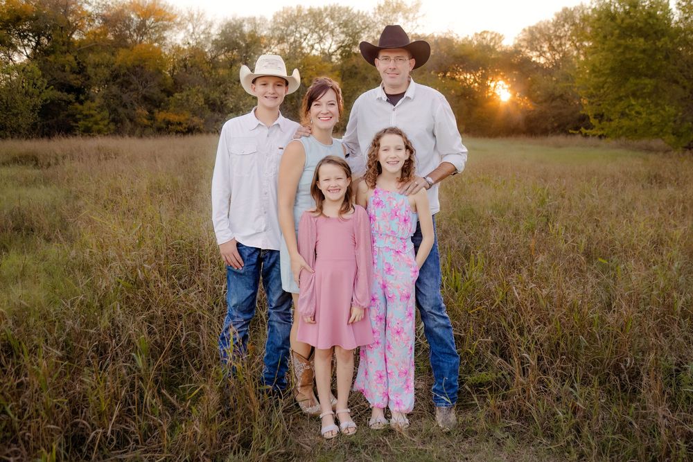 Family of five posing in golden hour sunlight in a field, wearing western attire including cowboy hats and pink dresses.