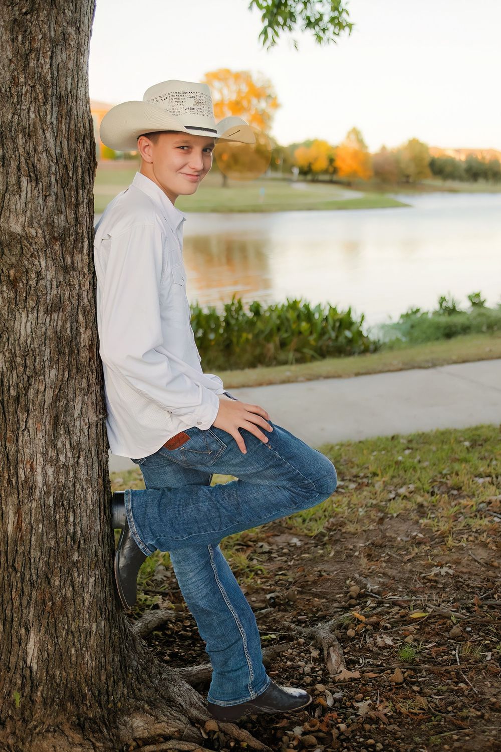 A young cowboy in a white shirt, jeans, and cowboy hat leans against a tree by a serene lake at sunset.