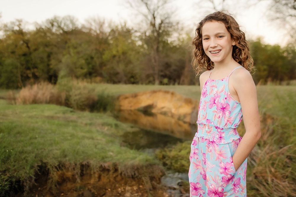 A smiling girl in a floral blue and pink jumpsuit poses outdoors near a creek in a lush green field.