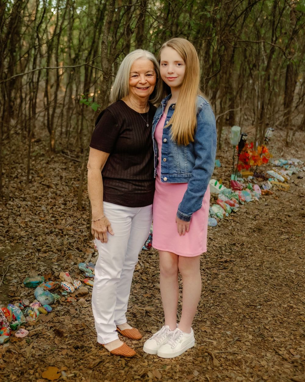Two women pose together in a wooded area with colorful decorations on the ground behind them.