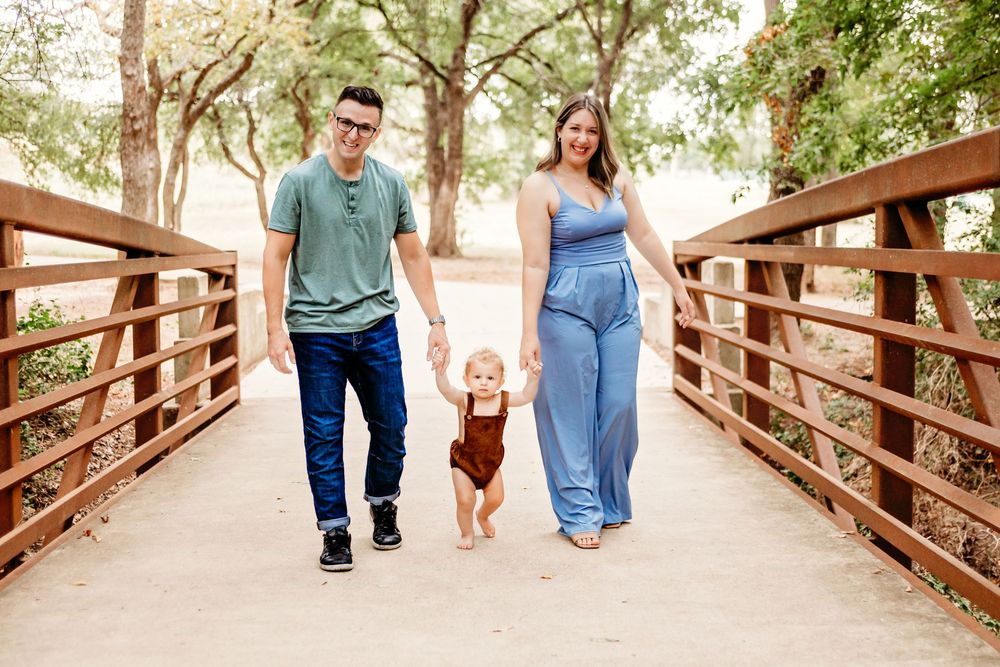 Family of three walking on wooden bridge, parents holding toddler's hands, surrounded by trees.