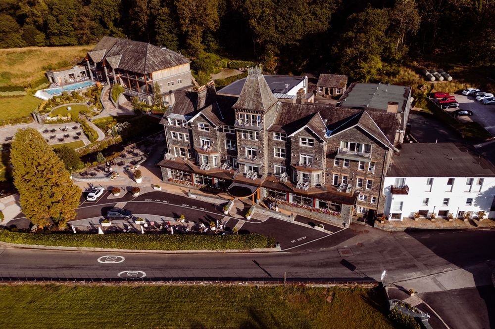 Aerial view of a large historic stone hotel surrounded by lush greenery and a winding road in the countryside.