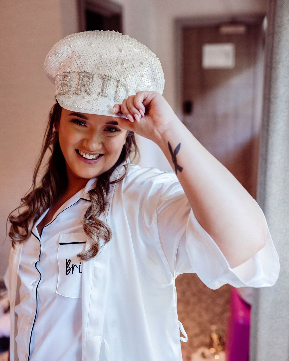 Happy bride wearing sequin BRIDE cap and white satin robe during wedding morning preparations.