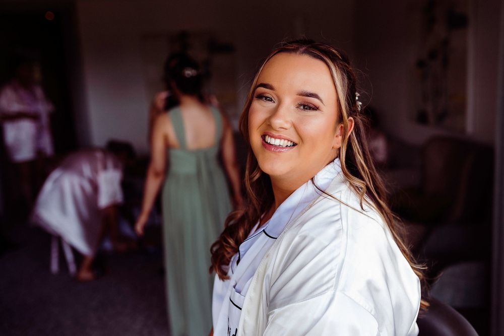 Smiling bride in white robe during wedding preparations with bridesmaids in green dresses in background.