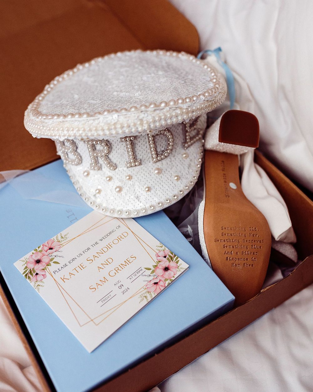 Wedding flat lay featuring pearl BRIDE cap, floral invitation, and bridal shoes in gift box.