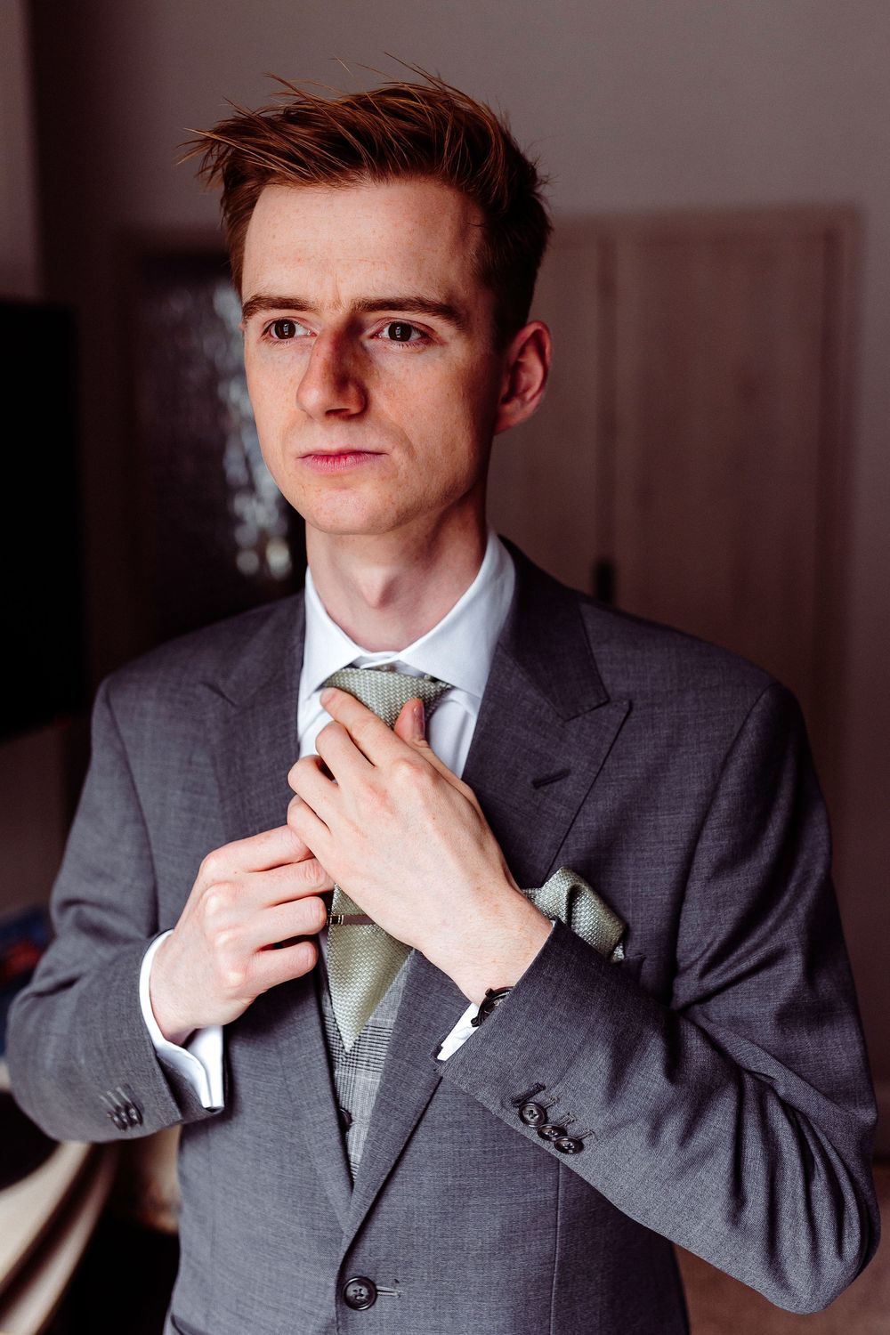 Elegant groom in grey three-piece suit adjusting his sage green tie before the wedding ceremony.