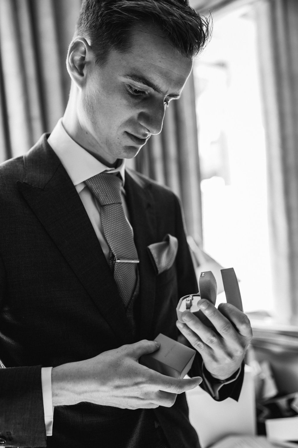 Black and white portrait of groom in suit examining wedding ring box before ceremony.