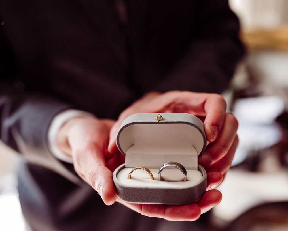 Person in dark suit holding open ring box with two wedding bands, one gold and one silver, on white velvet.