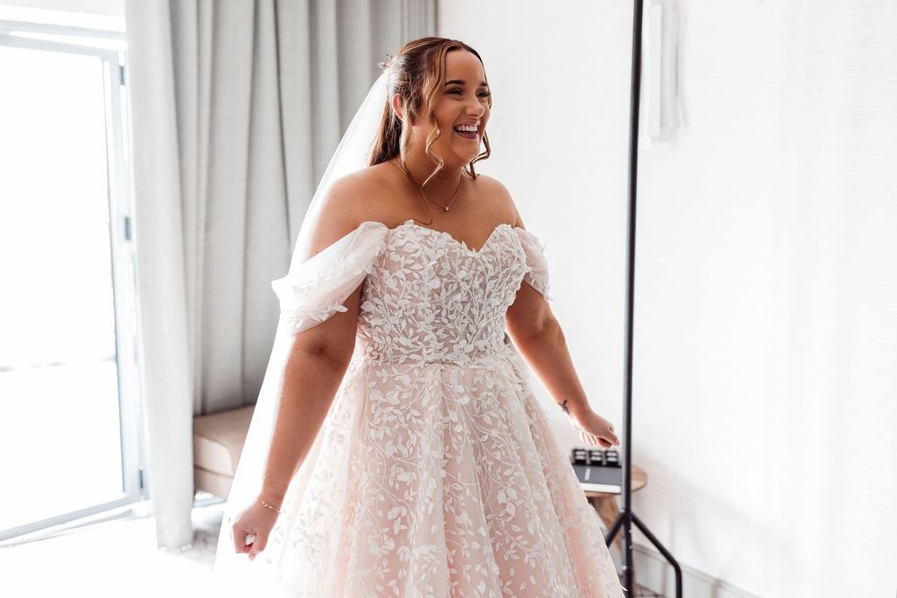 Smiling bride in off-shoulder lace ballgown wedding dress with veil, posing in bright fitting room.