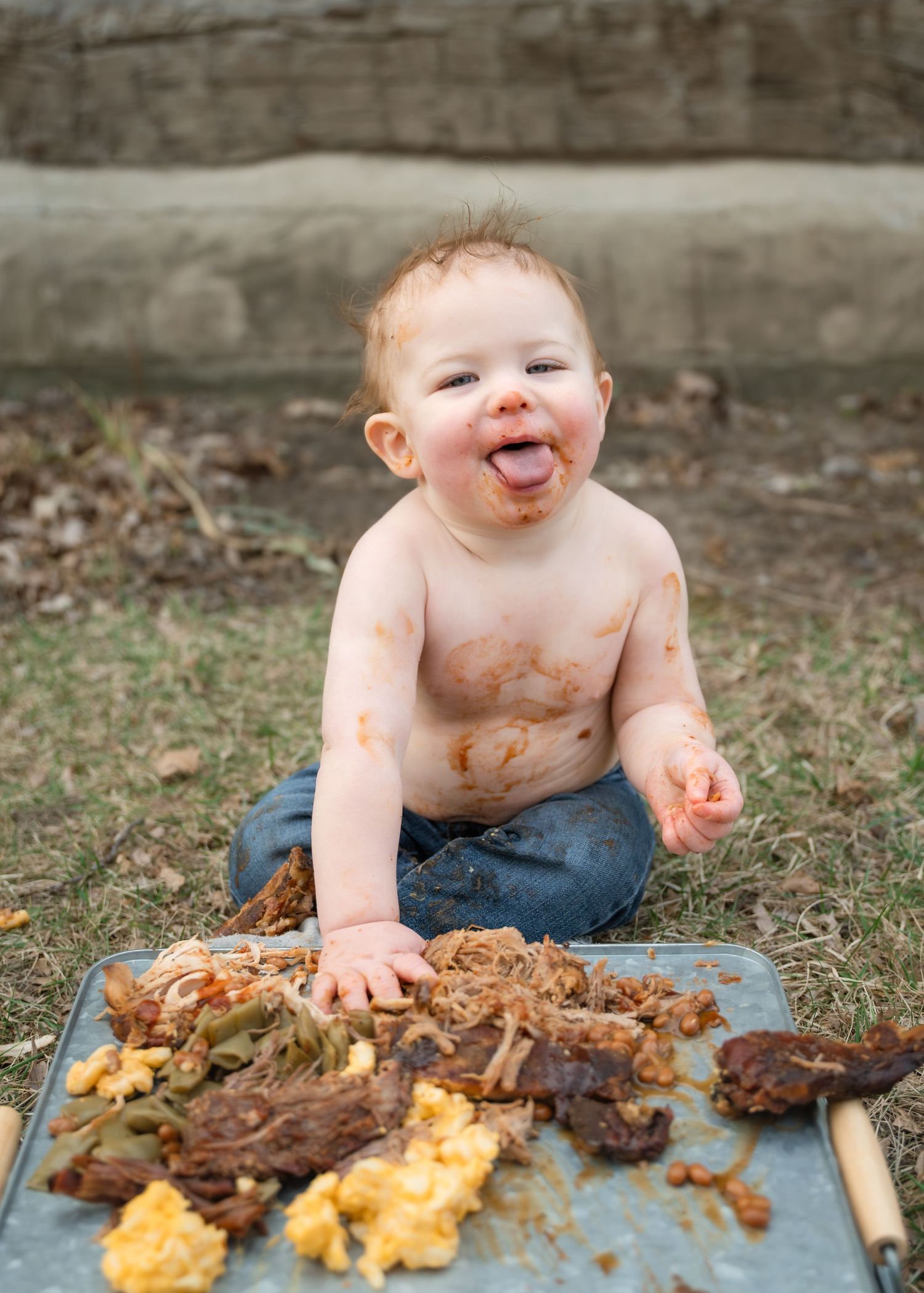 Messy baby with food smeared on face and chest sits outdoors on tray with scattered food, smiling happily.