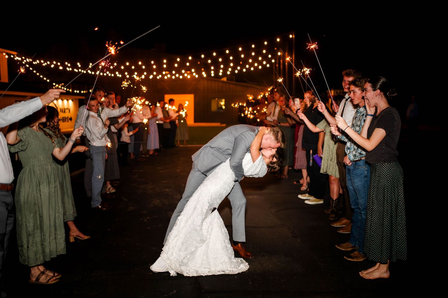 Bride and groom share a romantic dip kiss during sparkler send-off under string lights at night.