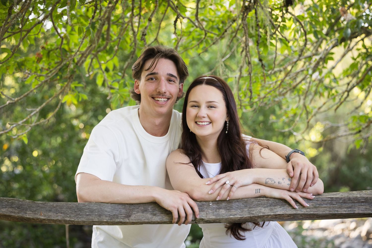 Smiling couple leaning on a wooden fence surrounded by lush green trees in an outdoor portrait.