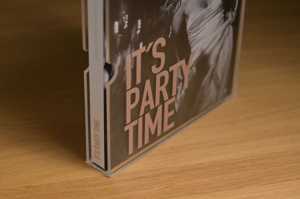 Close-up of Its Party Time photo book spine in transparent acrylic case on wood surface.