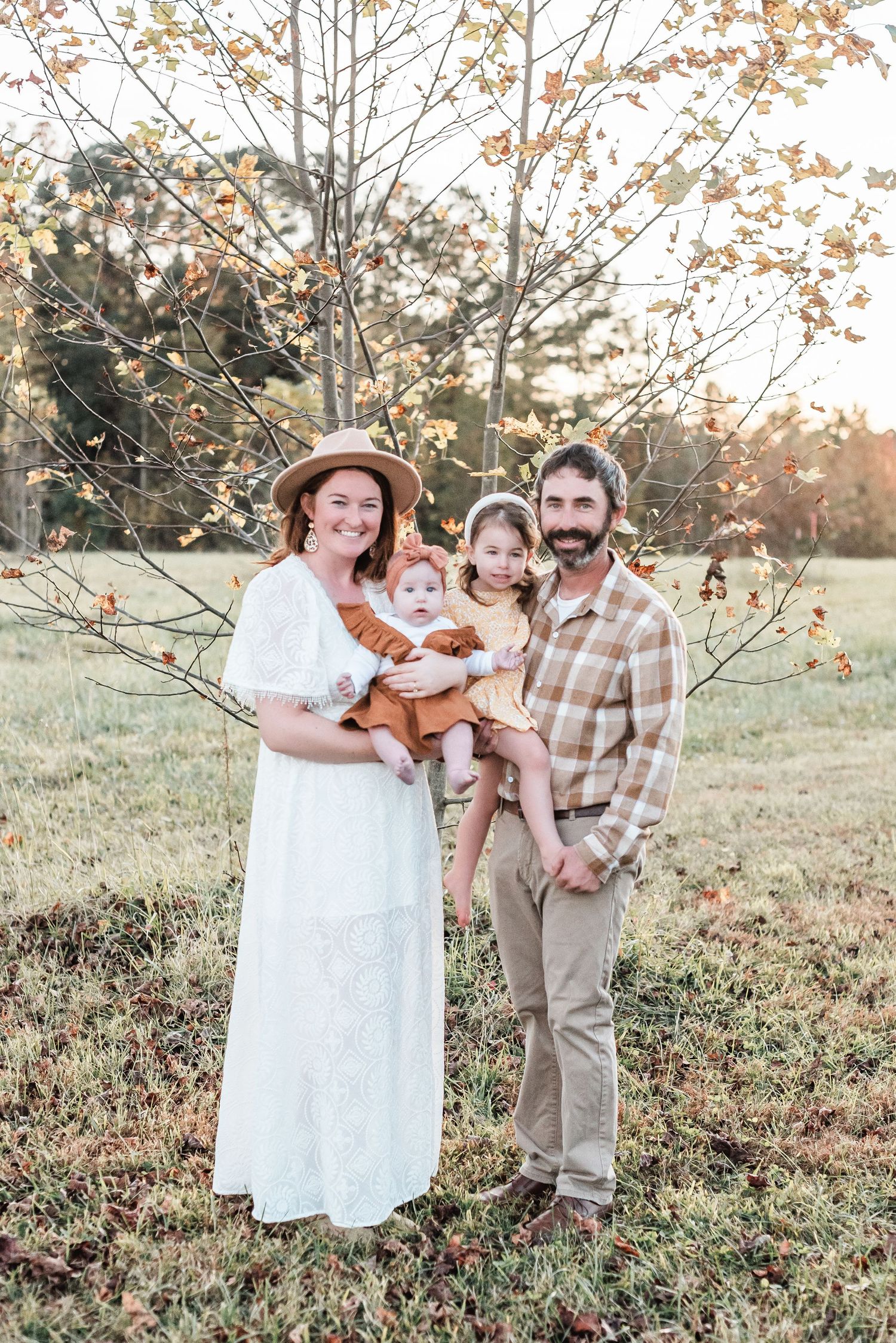 A family in white and plaid clothing poses together in a sunlit autumn field with golden foliage.