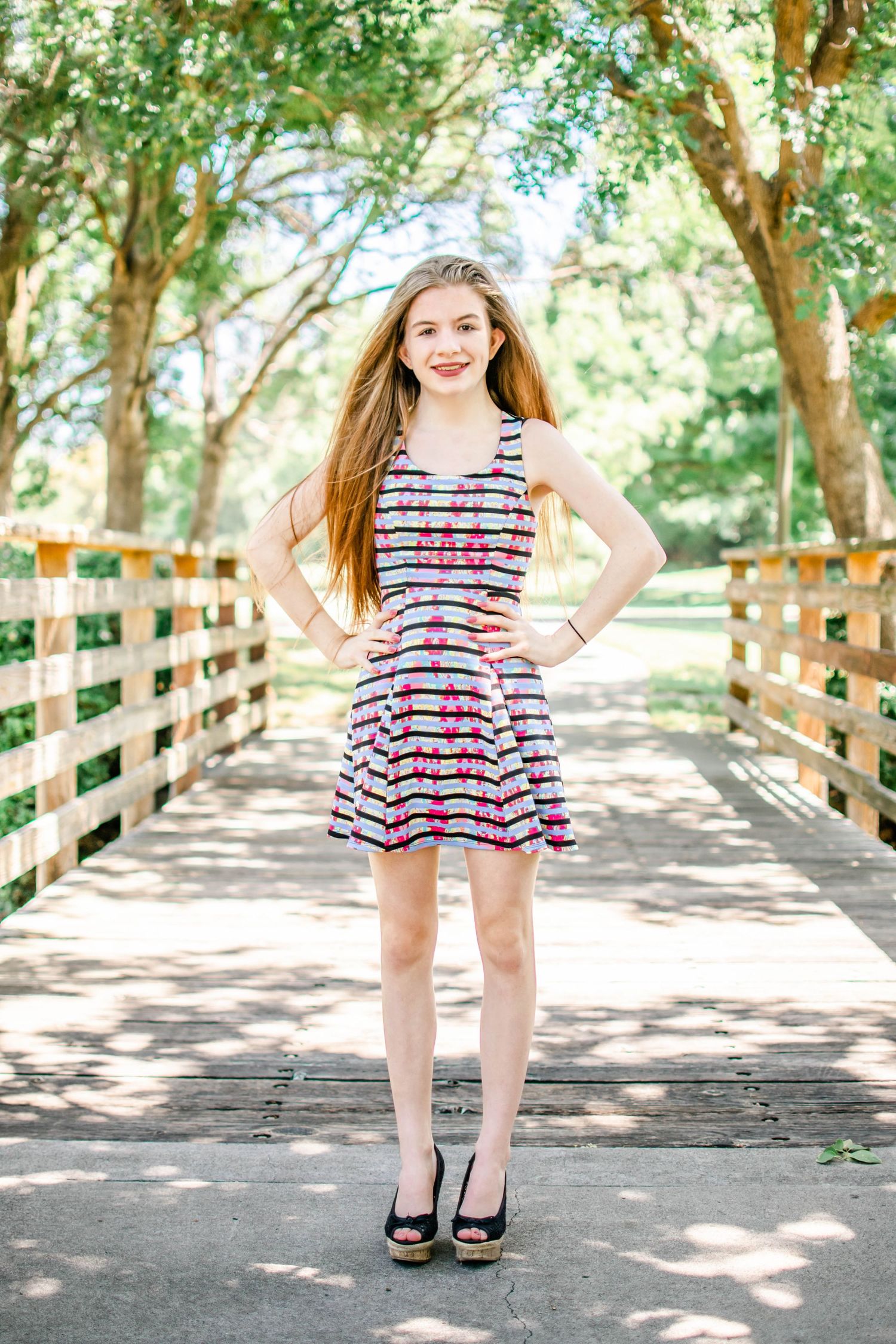 Young woman in floral dress posing on wooden bridge surrounded by lush green trees in a park setting.