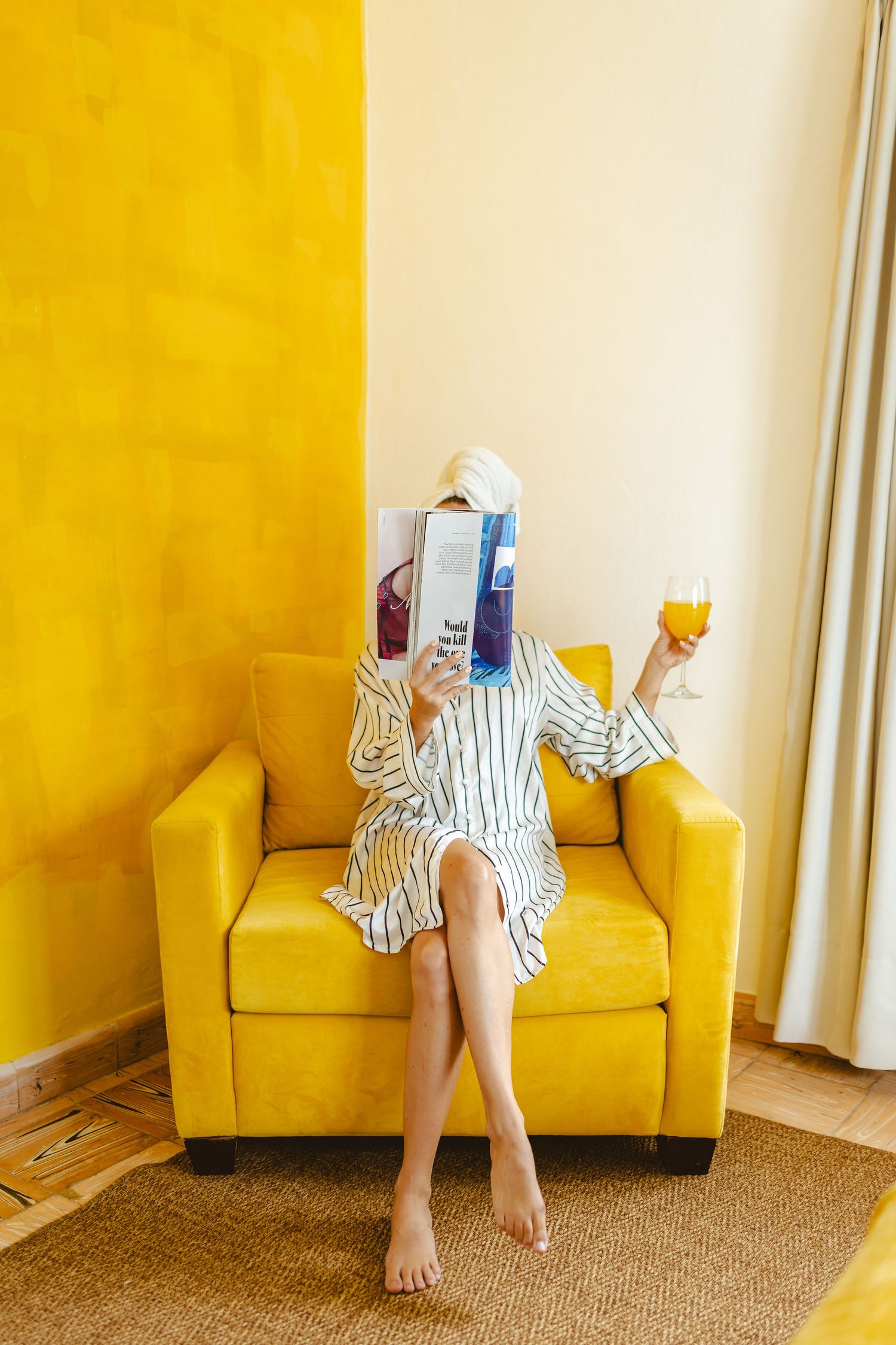 Woman in striped robe and towel turban reading magazines on toilet in modern white bathroom with patterned tile floor.