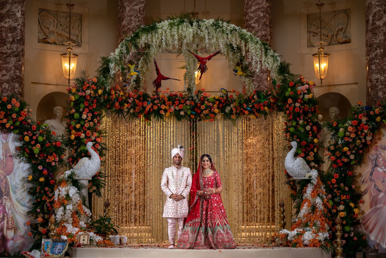 Indian bride and groom in traditional attire stand under floral arch with peacock decor at elegant wedding ceremony.