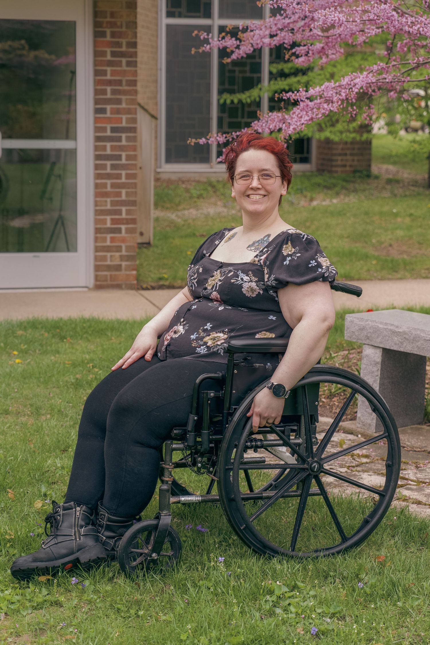 Smiling woman with red hair sitting in a wheelchair outdoors near a blooming pink tree and brick building.