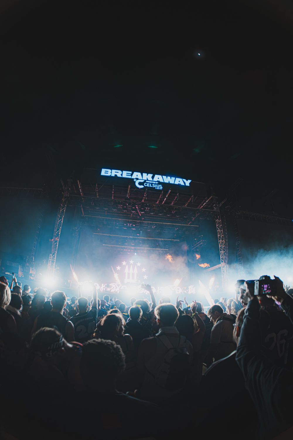 Energetic nighttime crowd at Breakaway festival facing a brightly lit smoke-filled stage with blue neon signage overhead.