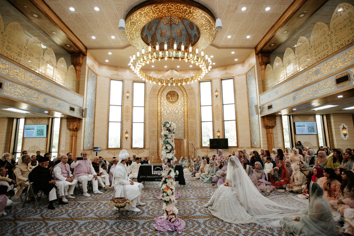 Muslim bride in white lehenga with veil sits during nikah ceremony in ornate mosque with gold calligraphy.