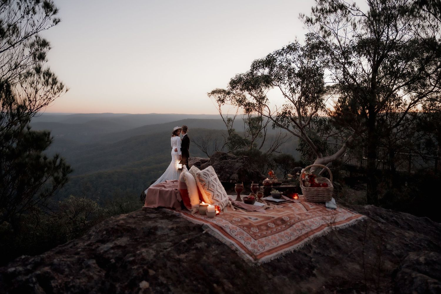 Couple standing on a rocky mountaintop at dusk with a romantic boho picnic setup and sweeping forest views.